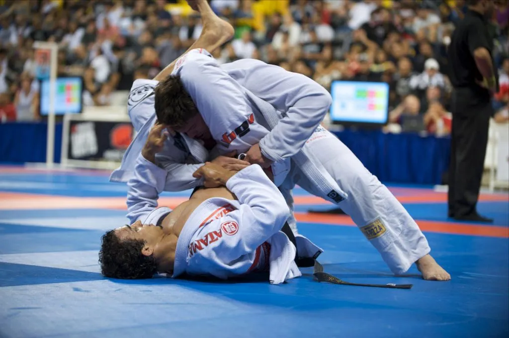 Two Brazilian jiu-jitsu competitors in white gis grappling at a tournament in front of a crowd