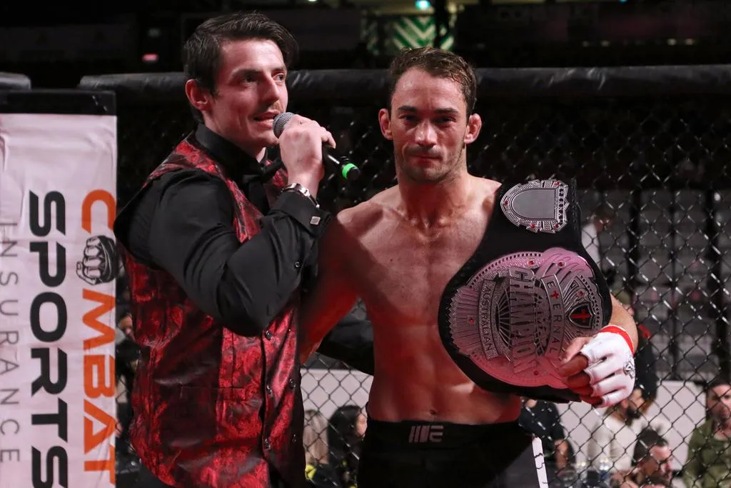 MMA fighter holding a championship belt while an announcer interviews him beside a Combat Sports Insurance banner