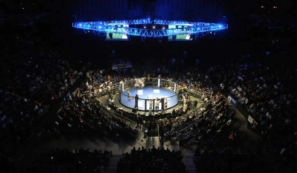 Boxing ring lit up in the centre of a dark arena packed with spectators