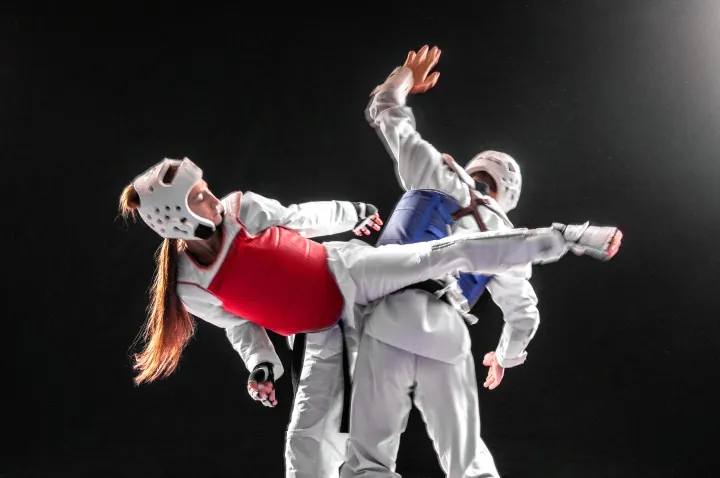 Two taekwondo athletes in headgear and chest protectors sparring, one throwing a high kick