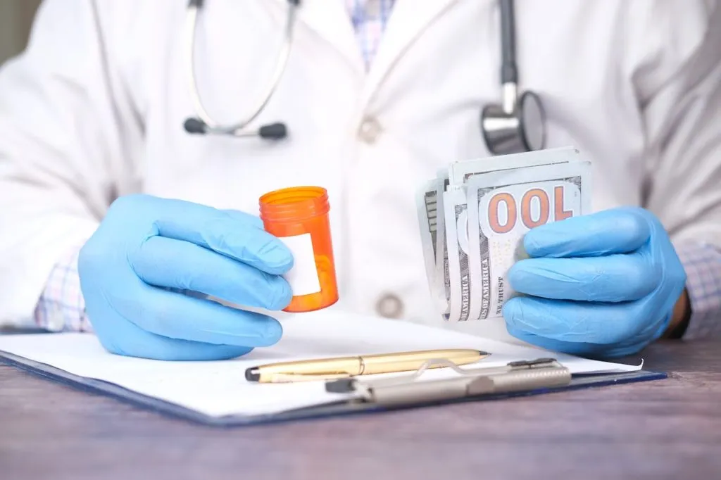 Doctor in blue gloves holding a pill bottle and a bundle of banknotes over a clipboard
