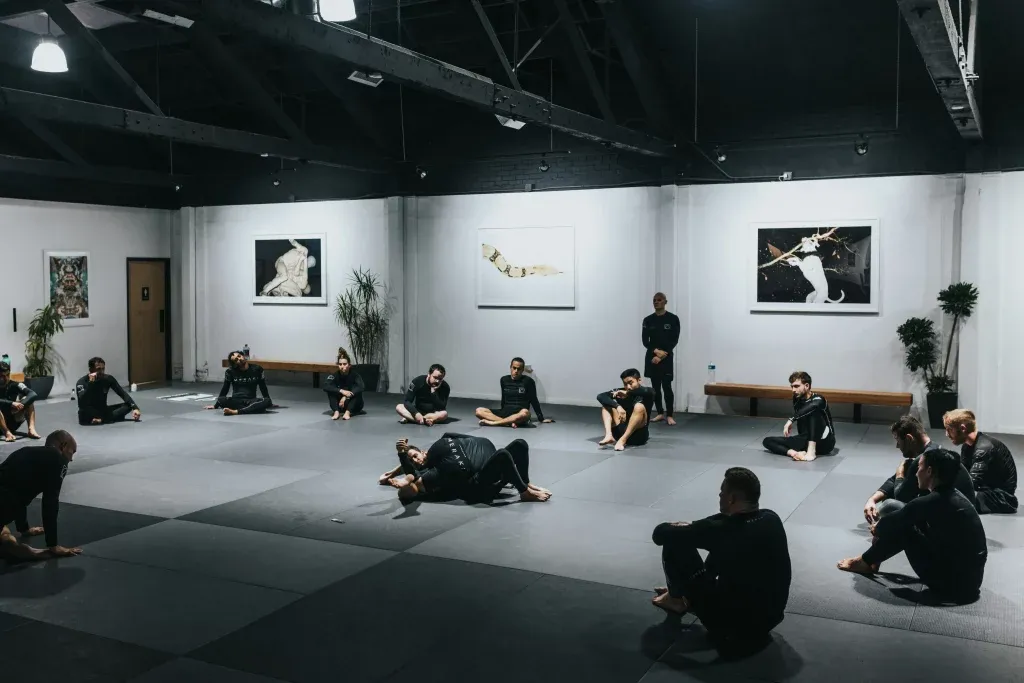 Martial arts students in black seated around the mat of a bright modern gym