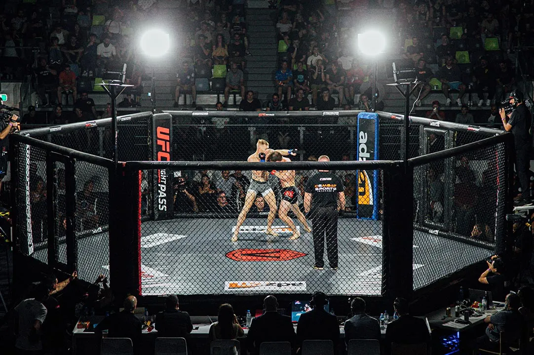 MMA cage in a packed arena with two fighters grappling and the referee close by