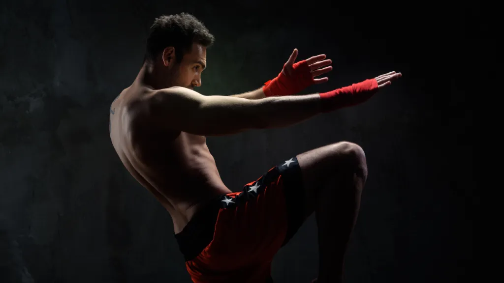 Muay Thai fighter in red shorts and hand wraps practising a knee strike against a dark background