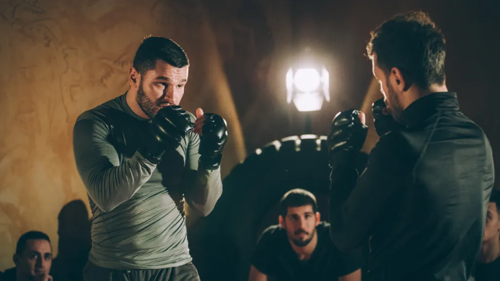 Two boxers in gloves squaring up in a dimly lit training room while others watch