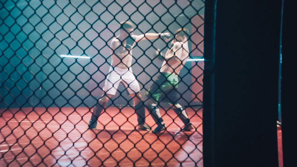Two fighters sparring in a cage under red and neon lights, seen through the chain-link