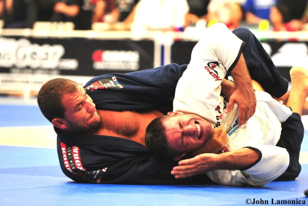 Two Brazilian jiu-jitsu competitors in dark gis grappling on the mat at a tournament