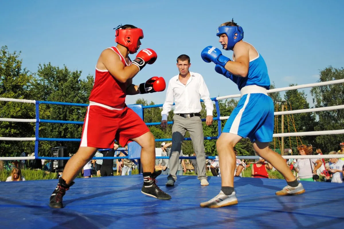 Two amateur boxers in red and blue sparring in an outdoor ring while the referee watches
