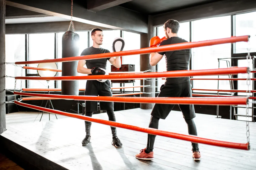 Coach holding pads for a boxer inside a red-roped ring in a bright gym