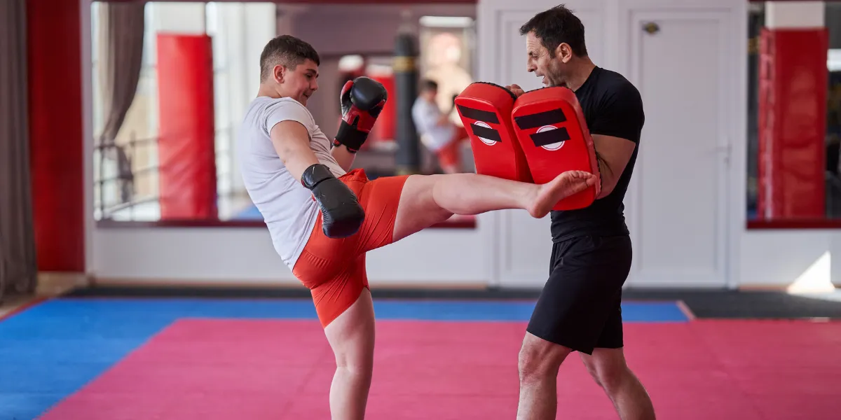 Kickboxer in red shorts kicking pads held by a coach in a gym
