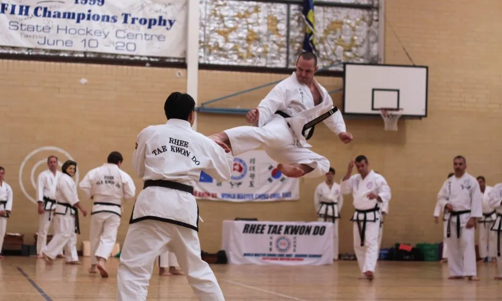 Taekwondo black belt performing a flying kick in a sports hall while students watch
