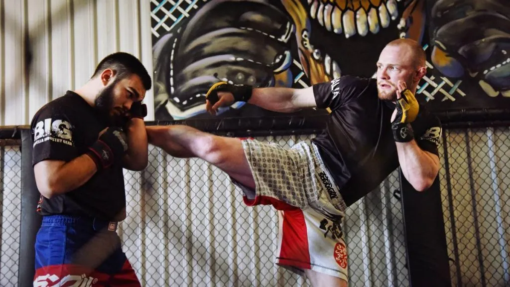 Fighter throwing a body kick at a training partner inside a gym cage