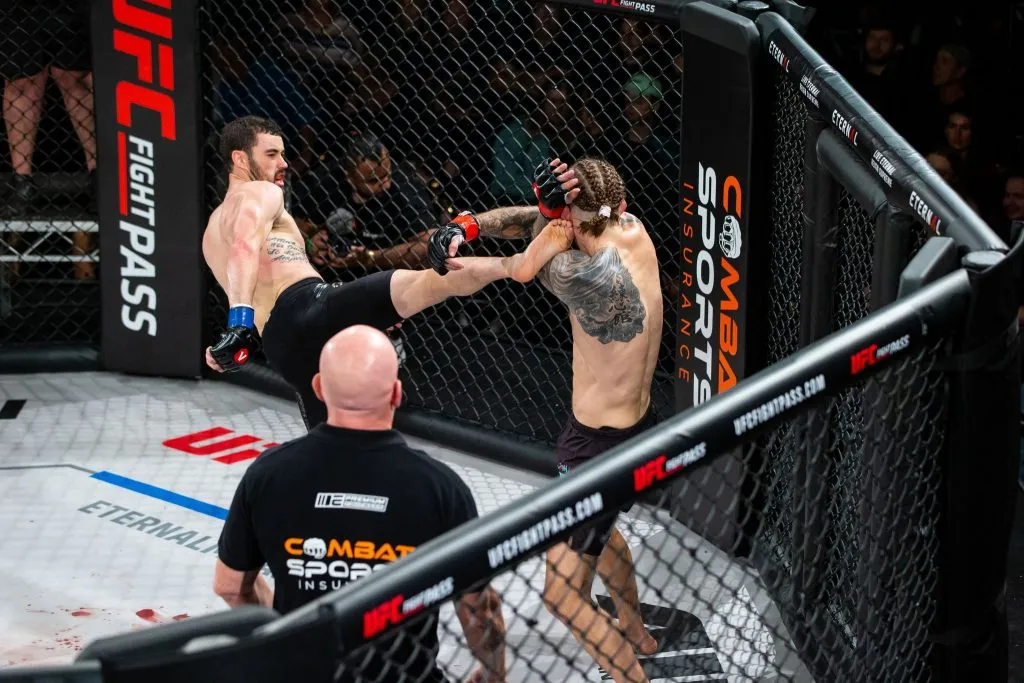 MMA fighter landing a head kick beside a Combat Sports Insurance cage banner while officials watch