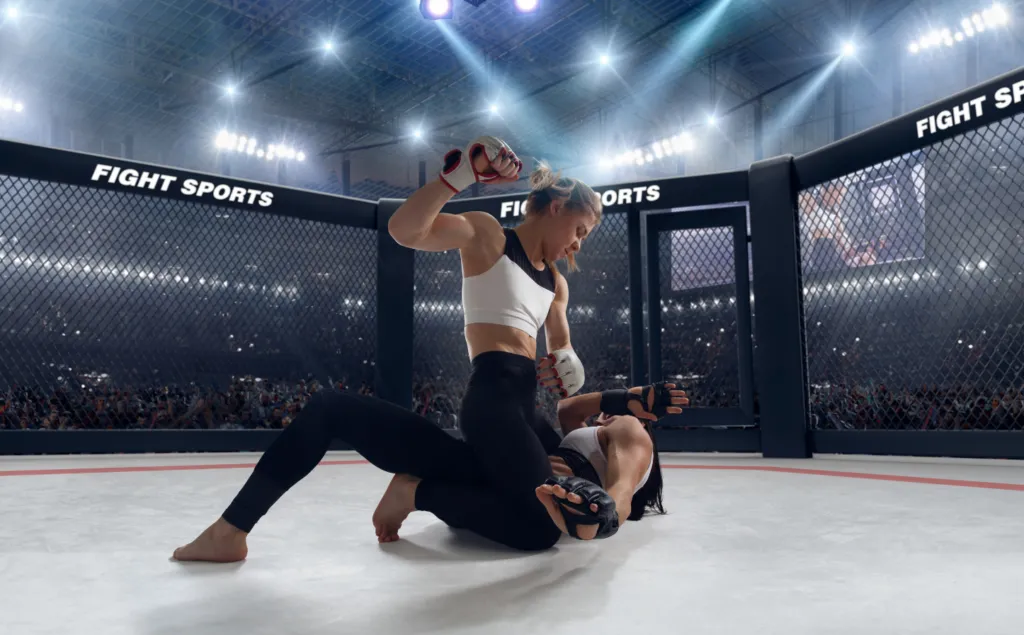 Female MMA fighter standing over a downed opponent in a brightly lit arena cage