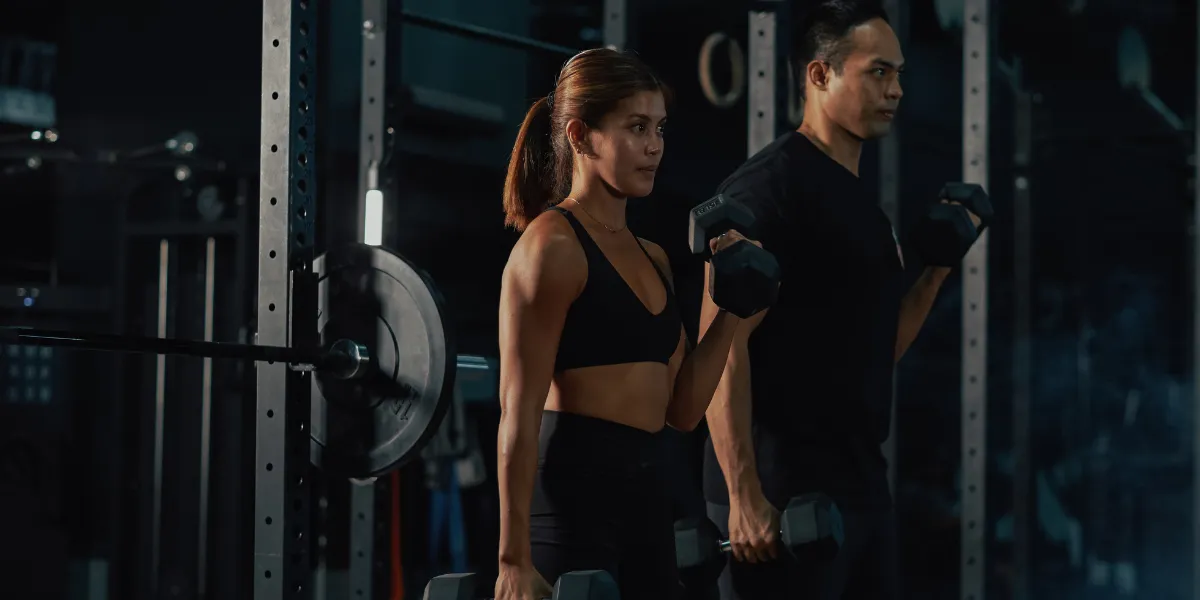 Woman and man holding dumbbells at their sides in a dark gym