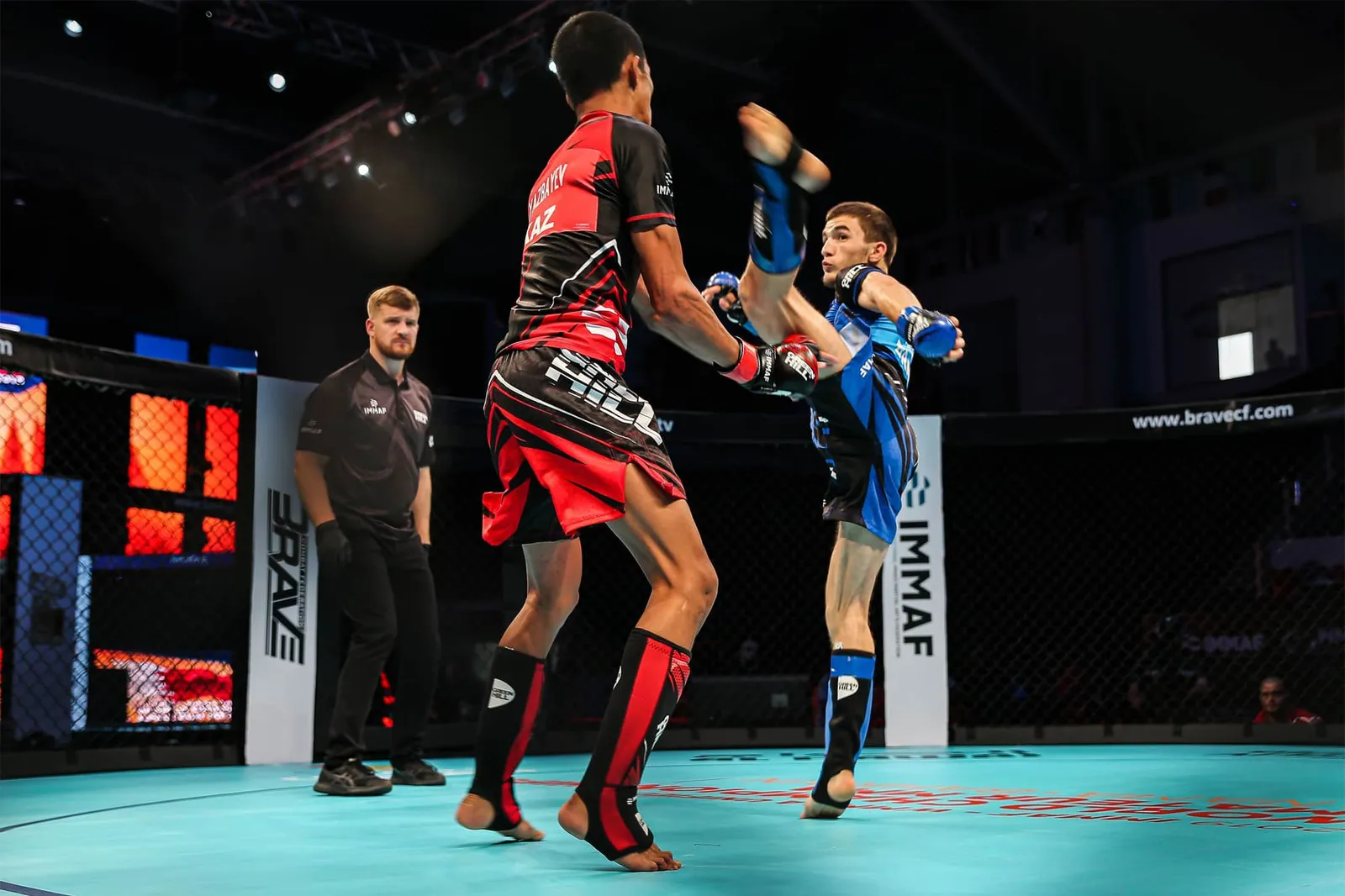 Two amateur MMA fighters in red and blue exchanging kicks in a cage while the referee watches