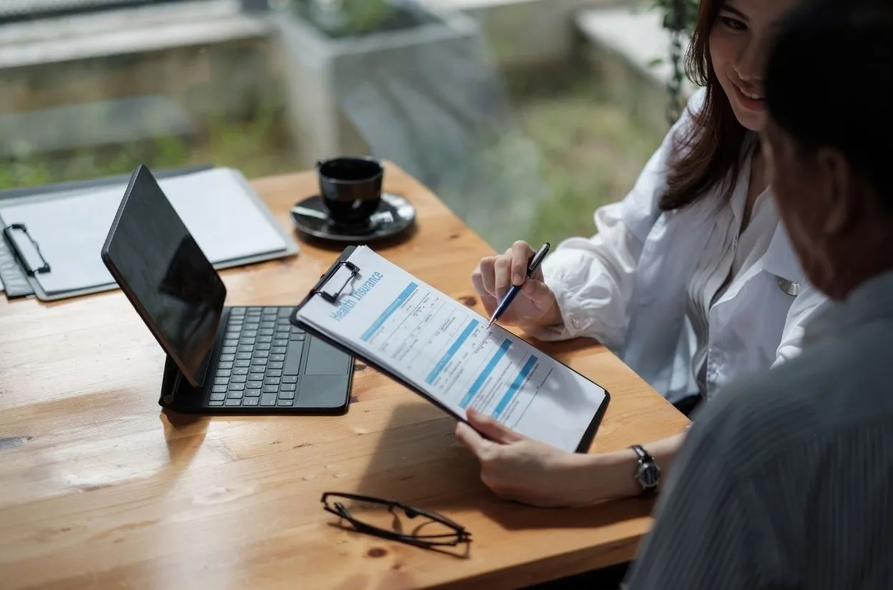 Adviser filling in a form on a clipboard while meeting a client at a wooden desk