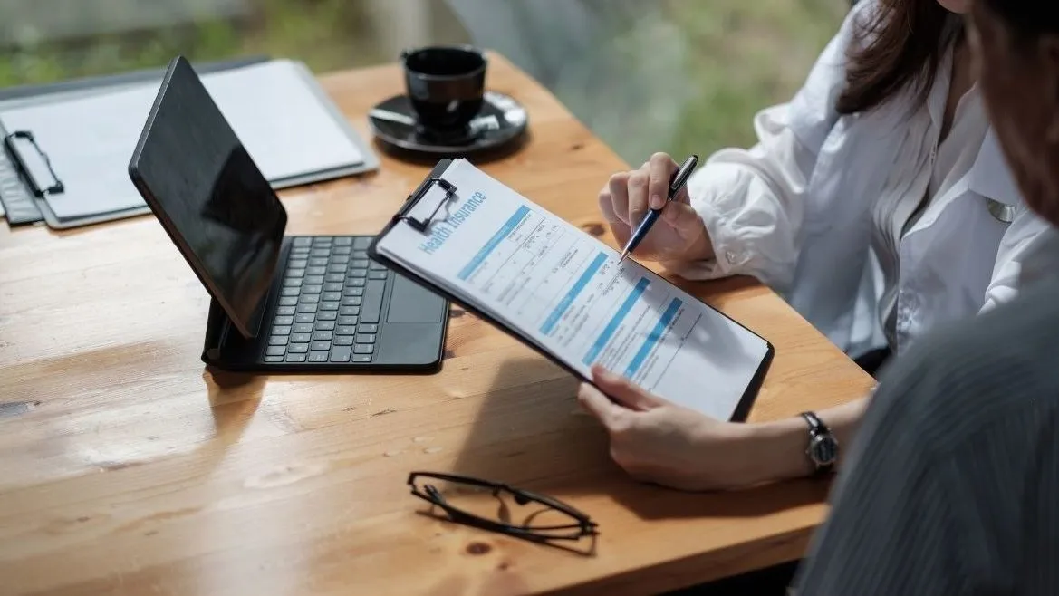 Person filling in a form on a clipboard at a wooden desk beside a laptop and coffee