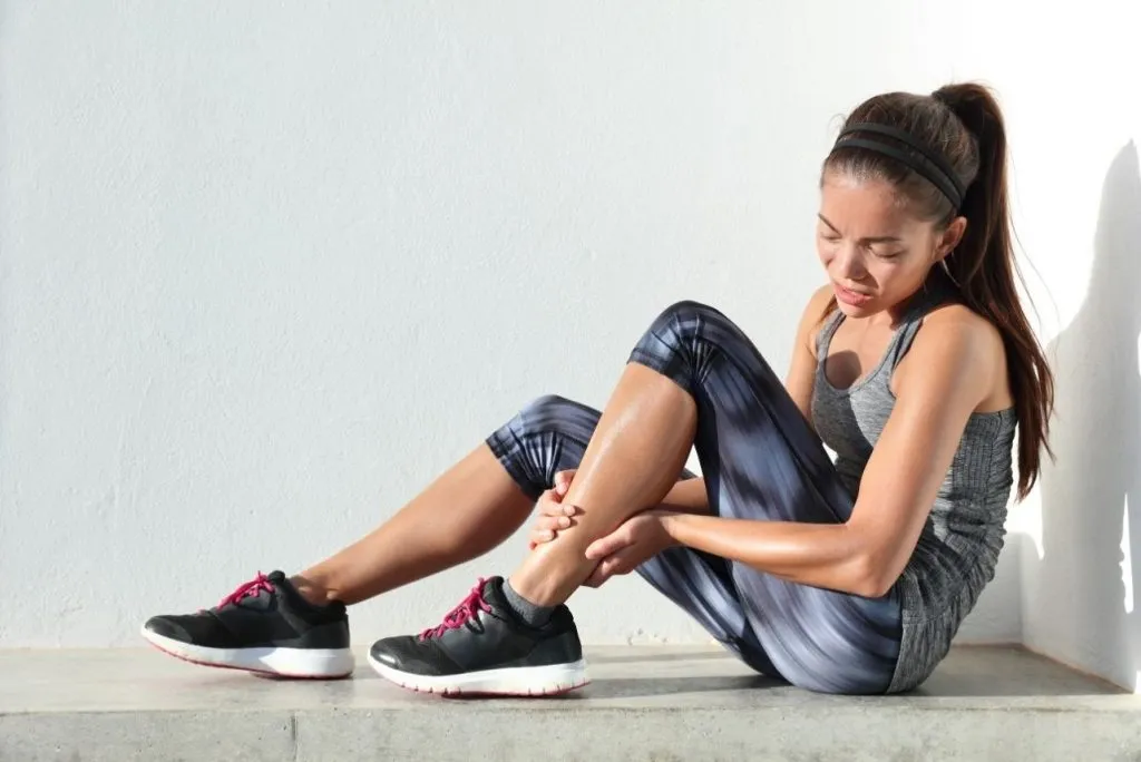Woman in running gear sitting against a wall and holding her injured ankle