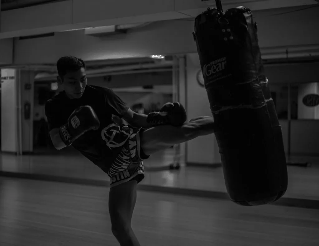 Black and white photo of a kickboxer kicking a heavy bag in a gym
