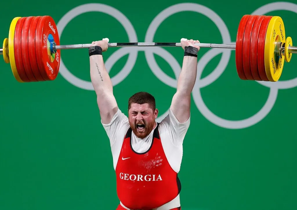 Weightlifter holding a loaded barbell overhead in front of the Olympic rings