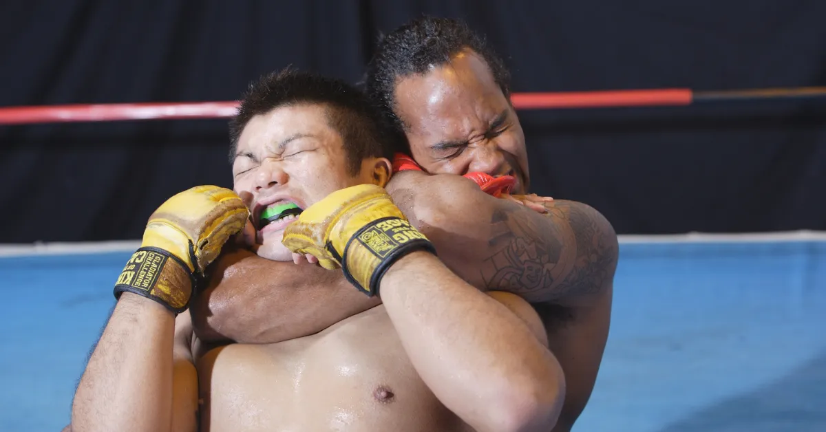 Two MMA fighters in a tight clinch, both grimacing, with one gripping the other's head