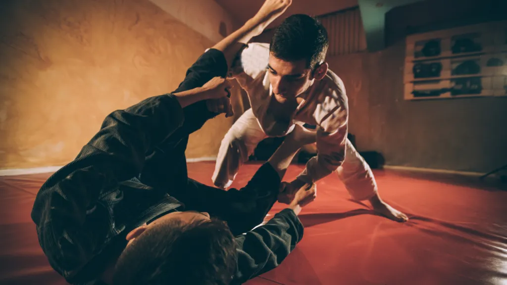 Two grapplers in gis training on a mat in a dimly lit gym