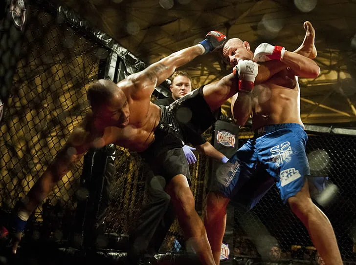 Two MMA fighters trading punches against the cage while the referee looks on