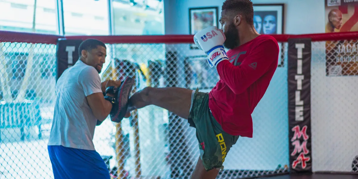 Fighter in a red shirt kicking pads held by a coach inside a cage at a gym