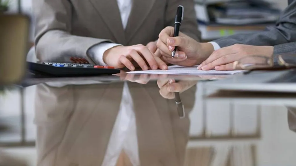 Person in a suit writing on paperwork next to a calculator