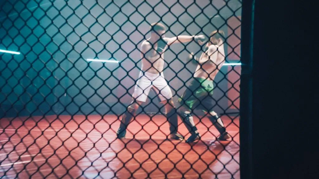 Two fighters sparring in a cage under neon lighting, seen through the chain-link fence