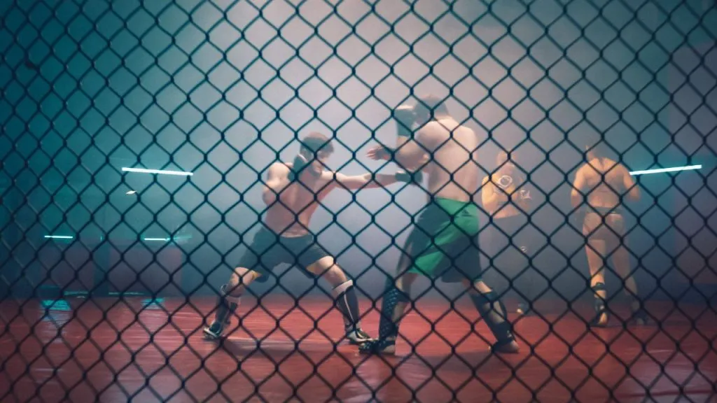 Two fighters sparring in a cage under neon lighting, seen through the chain-link fence