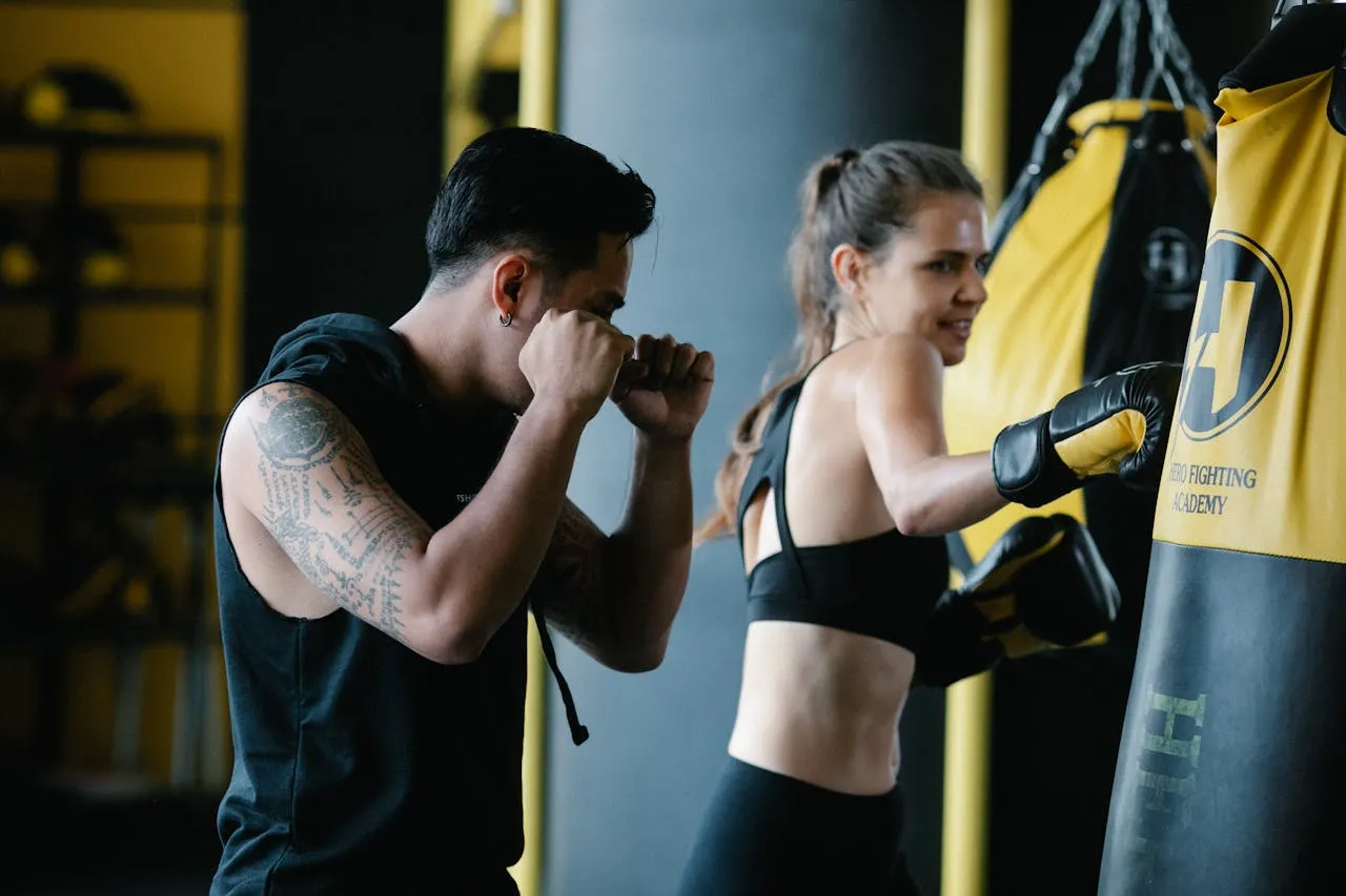 Coach in a guard stance beside a woman in gloves hitting a heavy bag in a gym