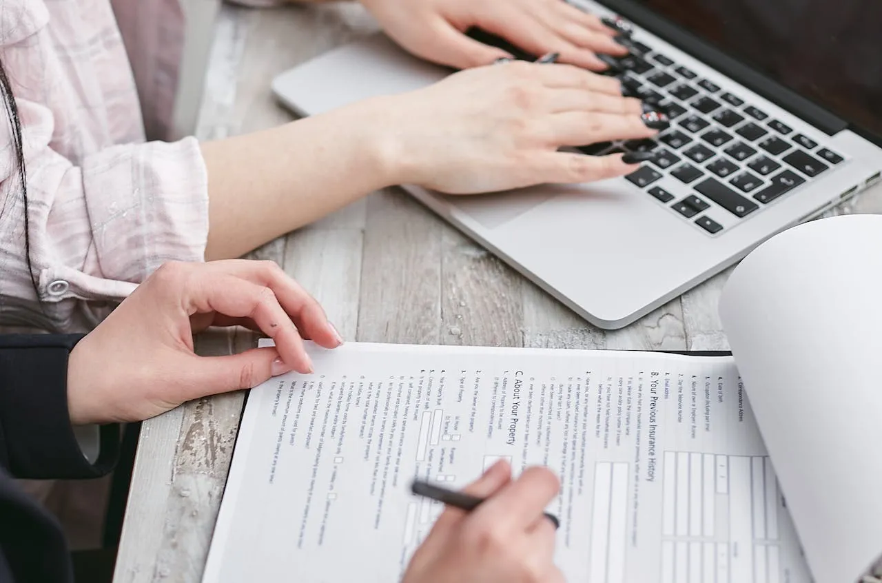 Person filling in a printed form at a desk beside a laptop