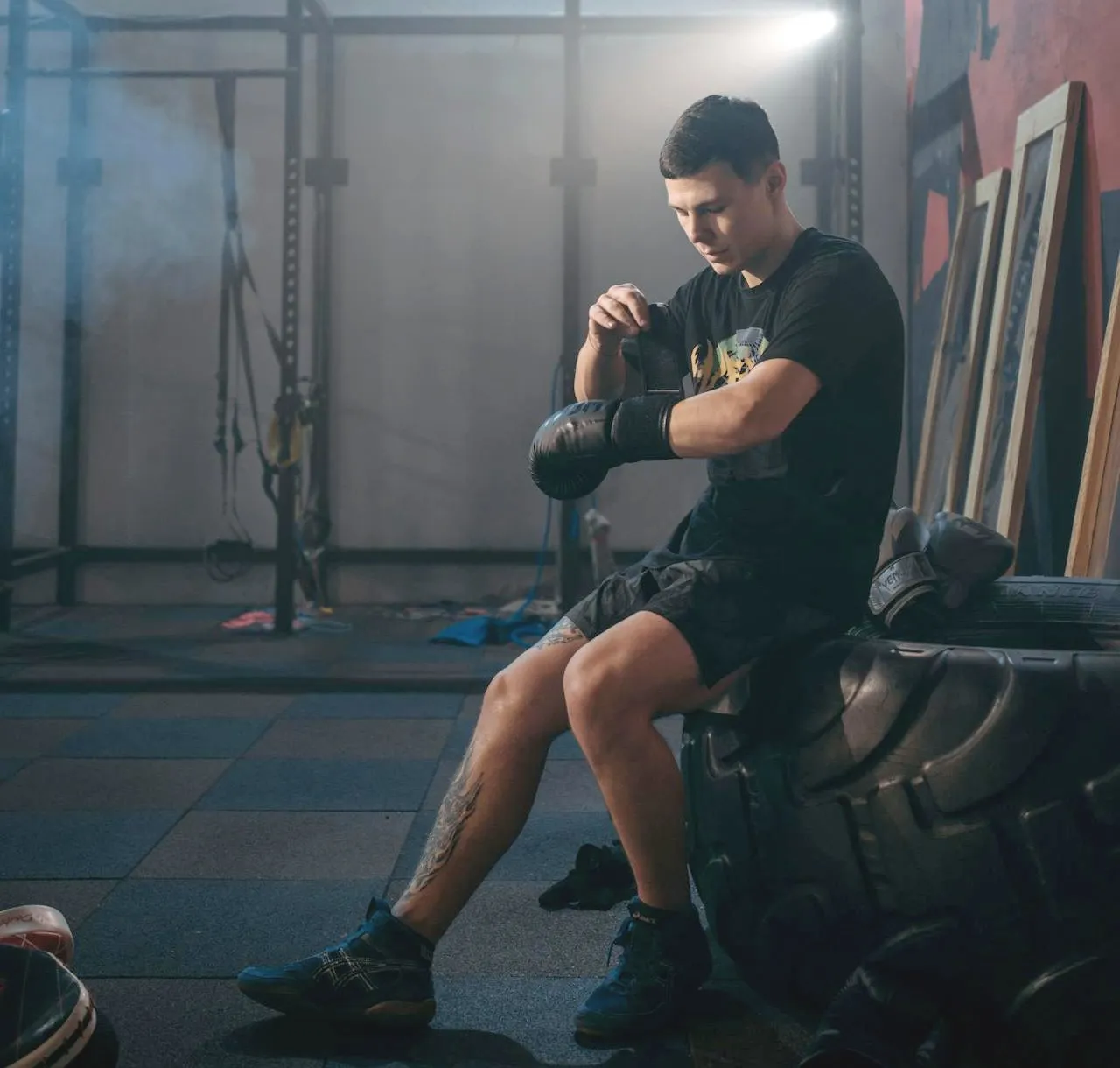 Fighter in gloves sitting on a tyre in a smoky gym adjusting his hand wraps