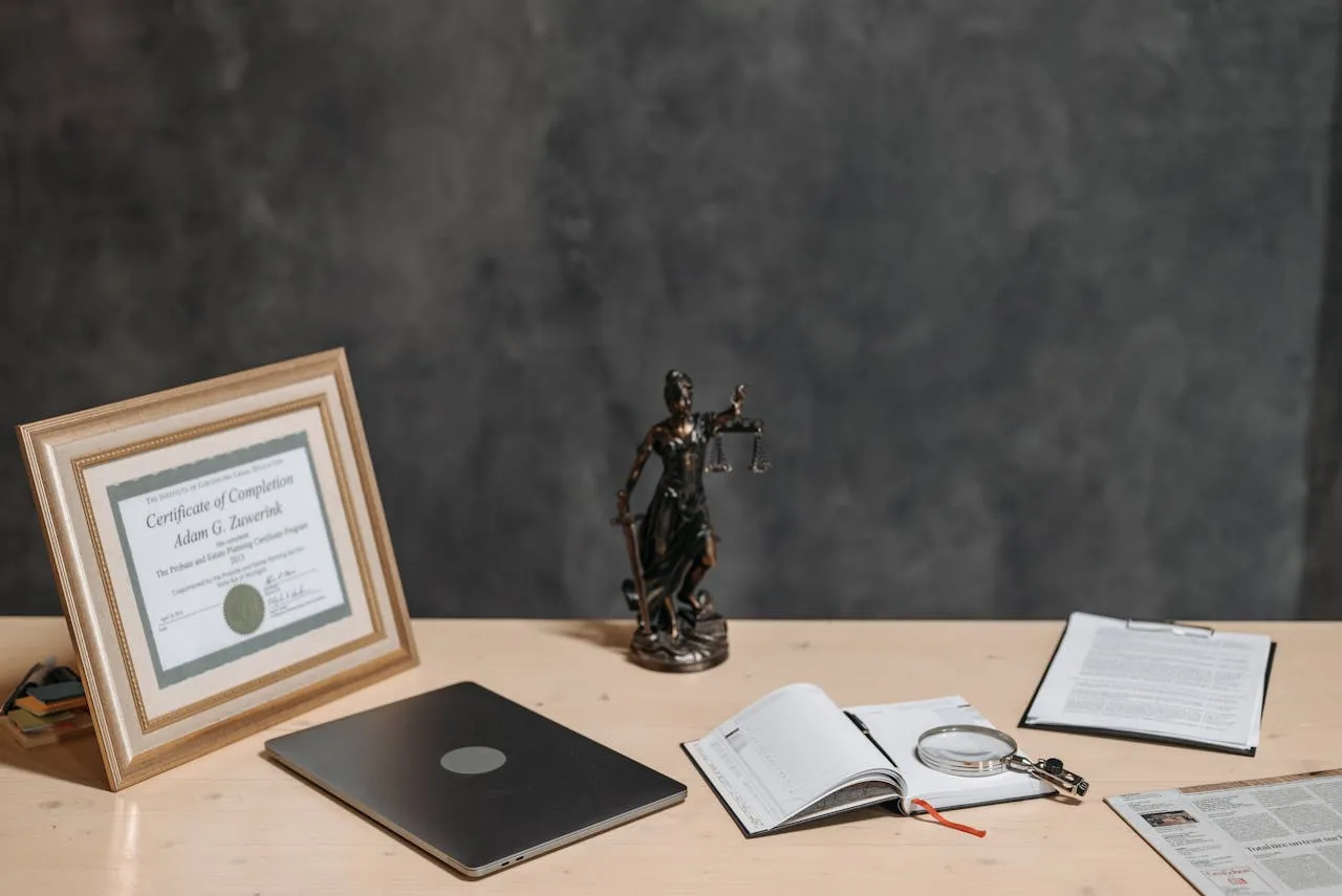 Framed certificate, a scales-of-justice figurine, a laptop and papers arranged on a desk