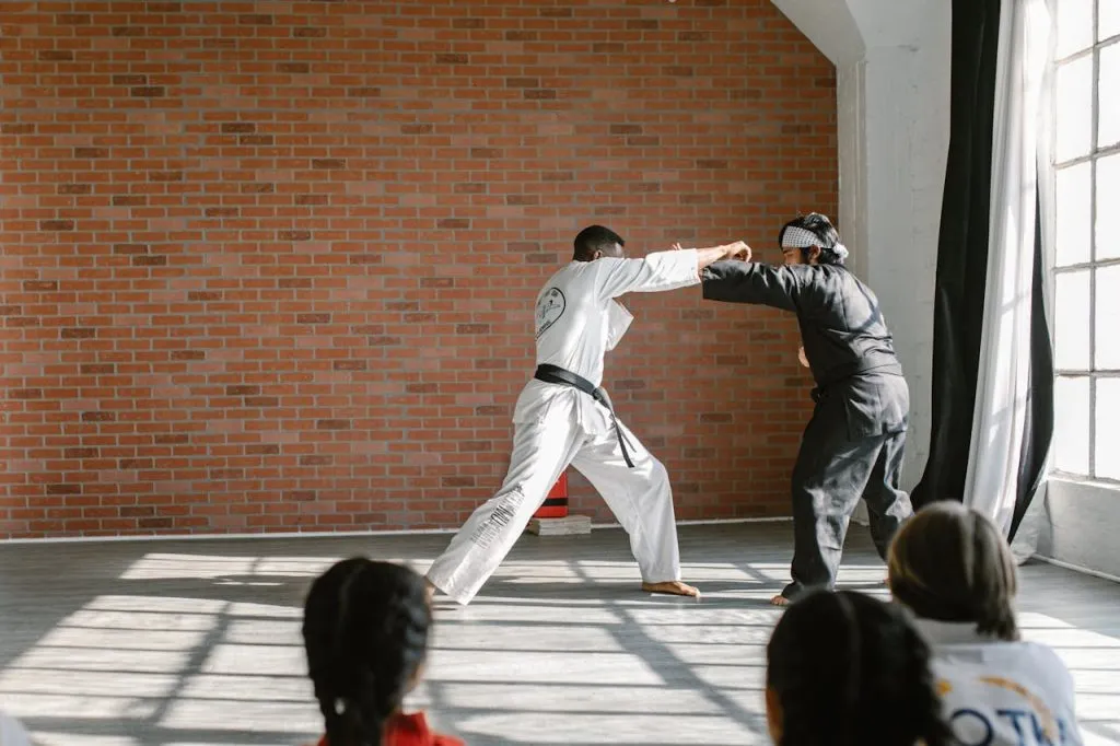 Instructor and student demonstrating a strike in a studio while children watch from the mat