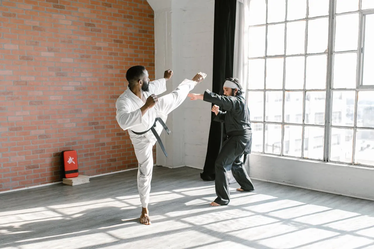Two martial artists sparring with kicks in a sunlit studio with brick walls