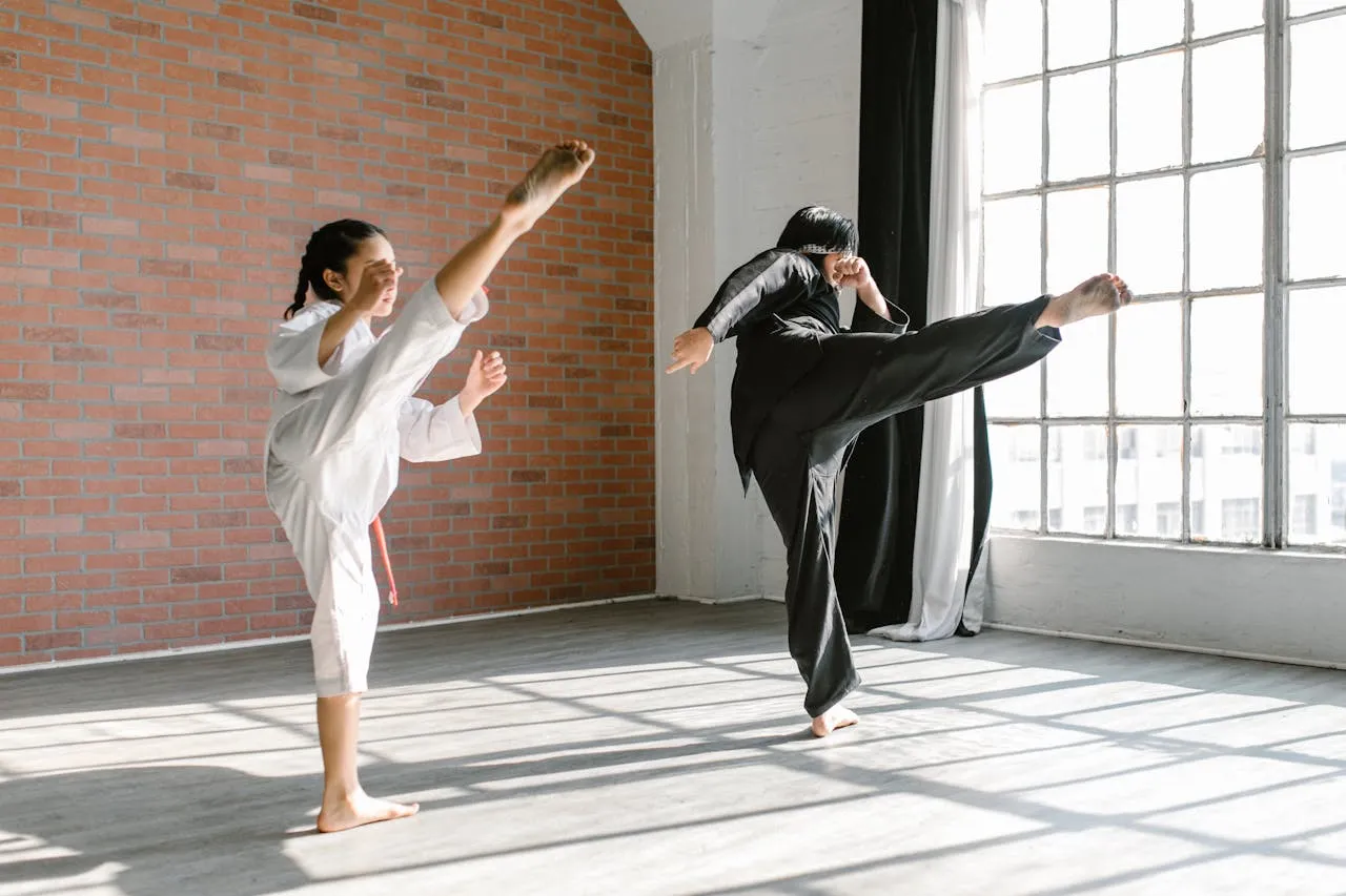 Two martial artists in black and white uniforms practising high kicks in a sunlit studio