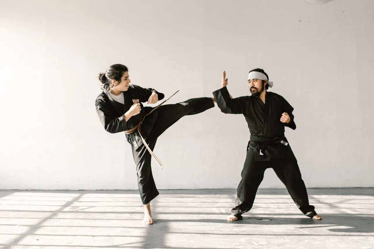 Two martial artists in black uniforms practising a kick and block in a bright studio