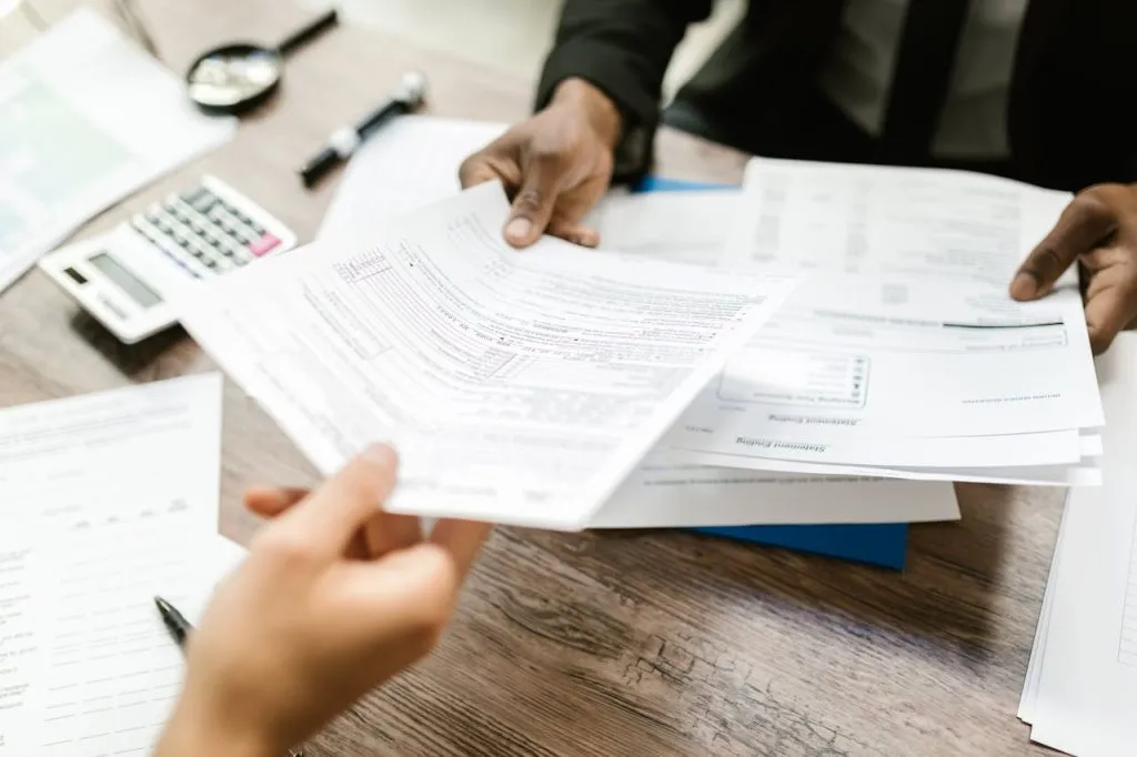 Two people passing insurance paperwork across a desk with a calculator
