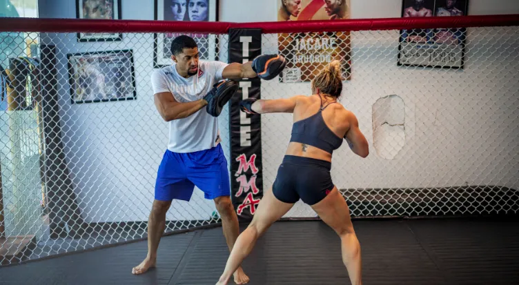 Coach holding pads for a female fighter inside an MMA cage at a gym