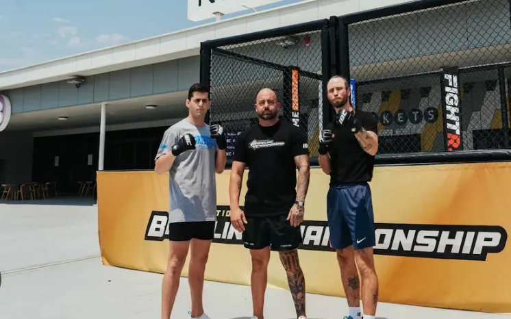 Three men in MMA gloves standing in front of an outdoor cage with a championship banner