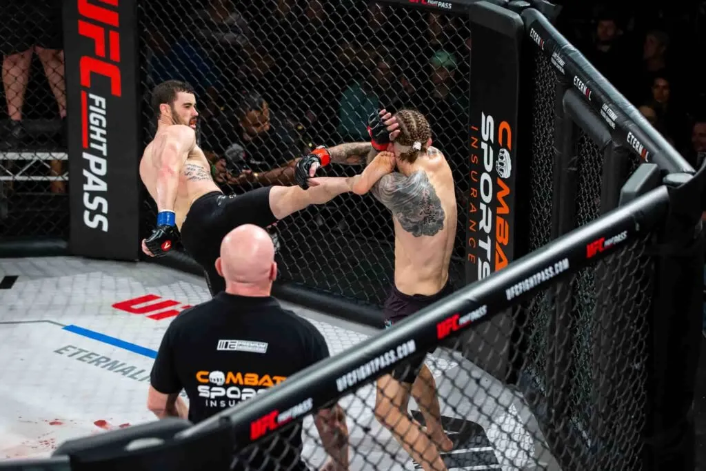 MMA fighter landing a head kick beside a Combat Sports Insurance cage banner while officials watch
