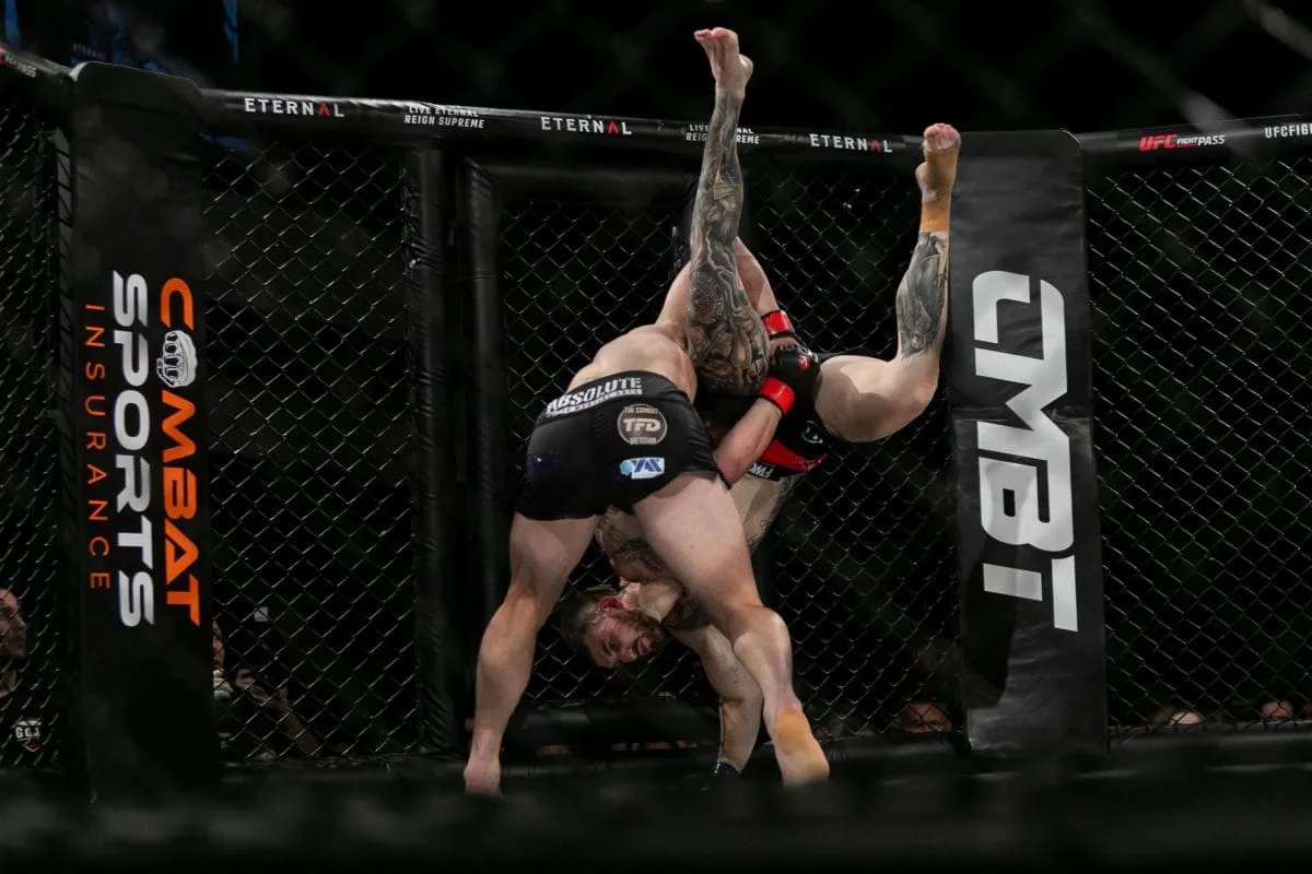 MMA fighter hoisting an opponent upside down for a slam next to a Combat Sports Insurance cage banner