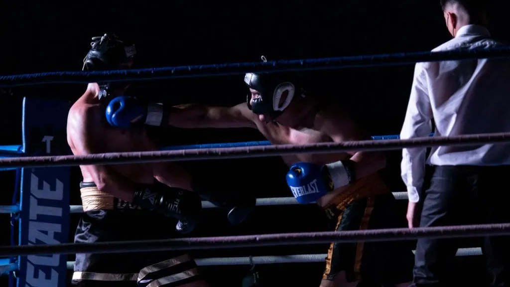 Two boxers in headgear exchanging punches in a dimly lit ring while the referee watches