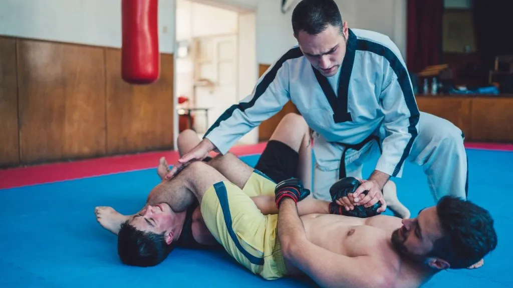 Jiu-jitsu instructor coaching two students practising a leg lock on blue mats