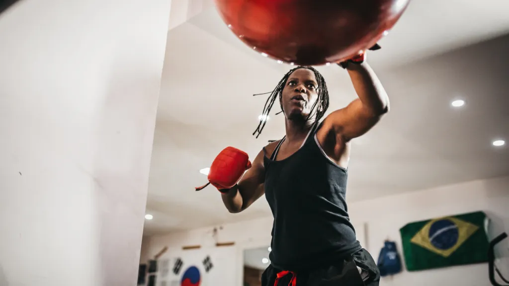 Woman in boxing gloves training with a medicine ball beneath a Brazilian flag in a gym