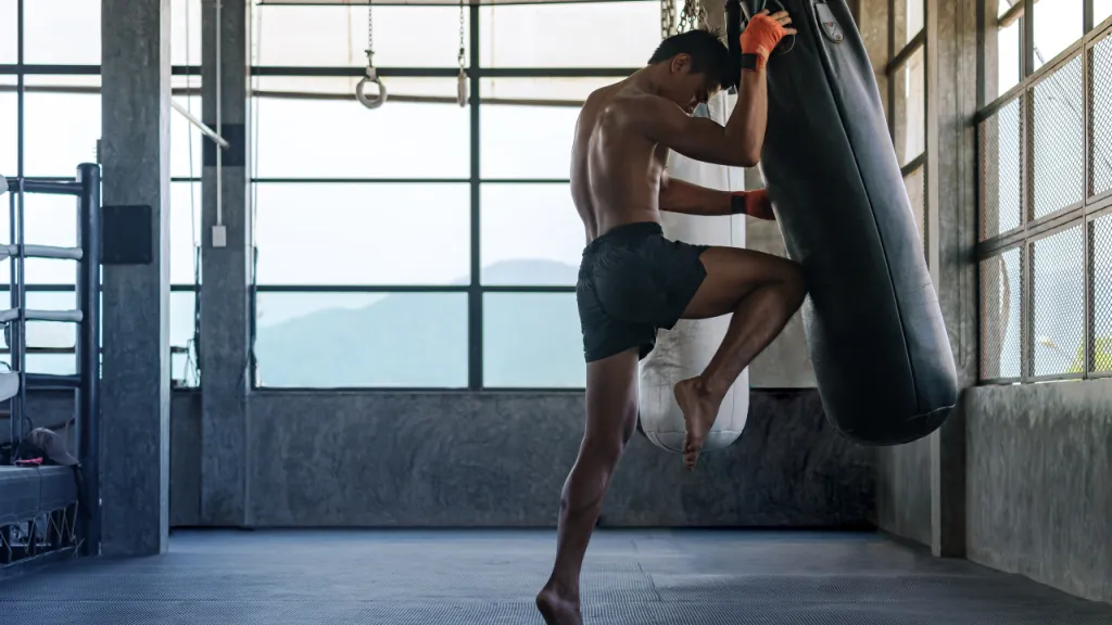 Fighter driving a knee into a heavy bag in a gym with large industrial windows