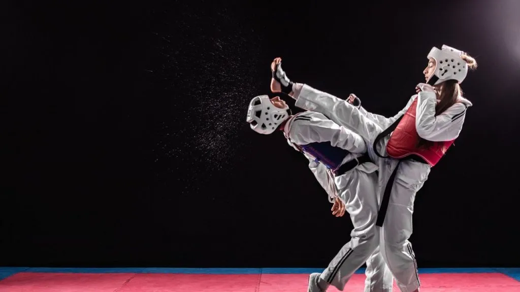 Two taekwondo athletes in headgear sparring, one landing a high kick